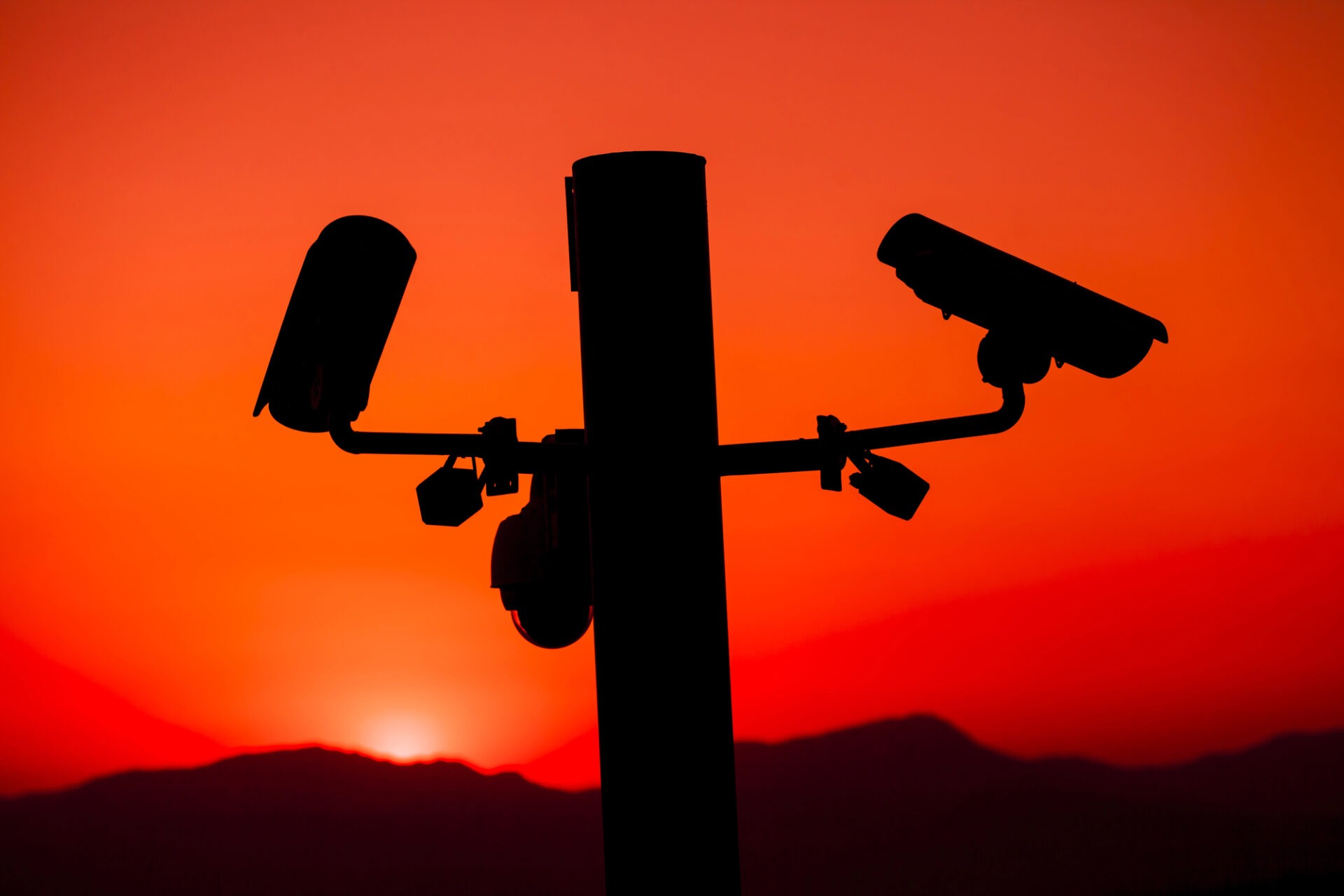 Silhouet van beveiligingscamera's op een paal tegen een felrode zonsondergang en donkere bergen.