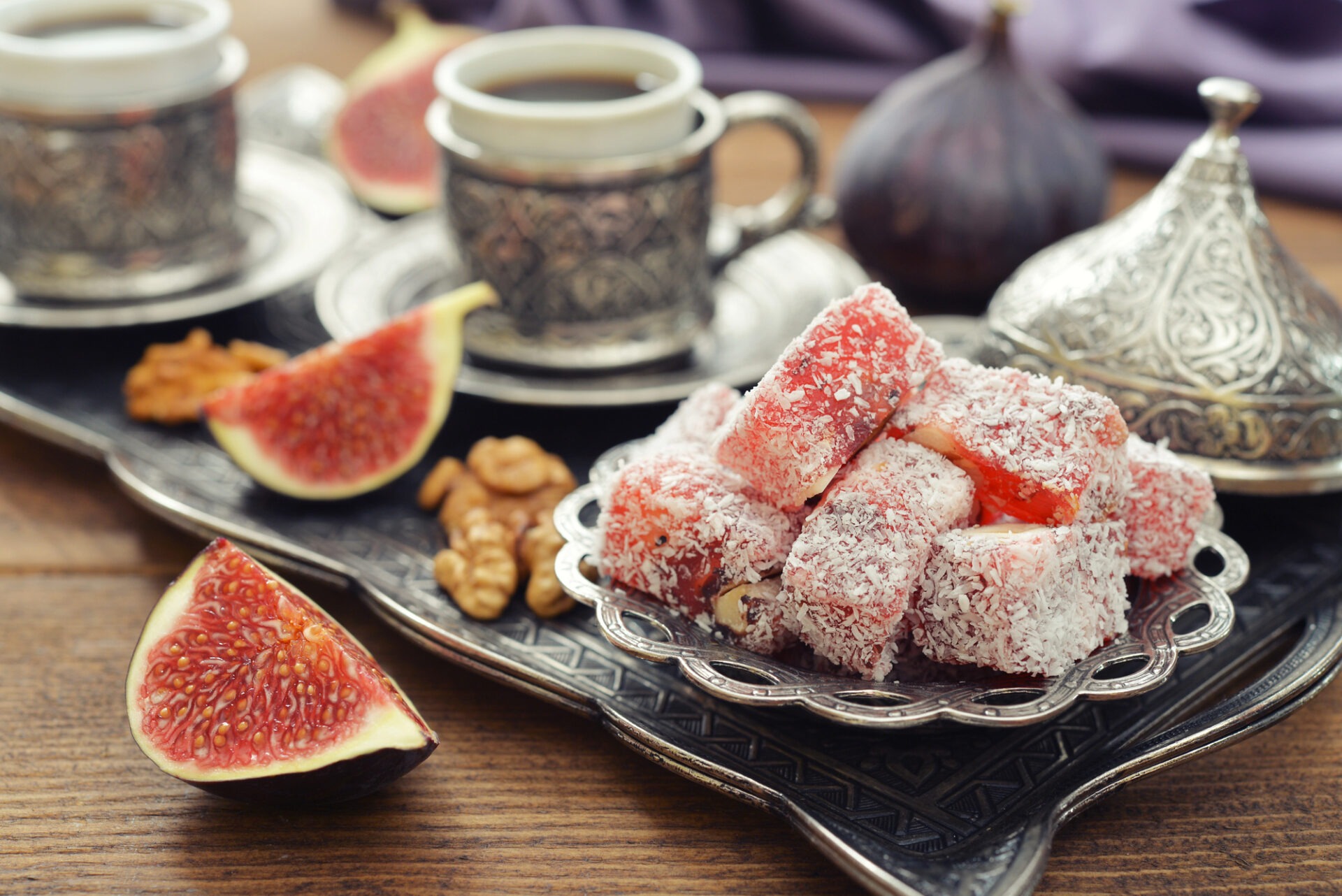 Zilveren schaal met roze Turks fruit, verse vijgen, walnoten en traditionele Turkse koffiekopjes op een houten tafel.